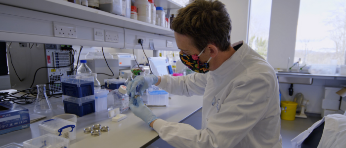 Man wearing face mask sat at a lab bench, wearing a lab coat and holding tools. 