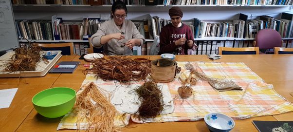 Wooden table with bundles of long plant fibres and bowls of water. Two women sat at table twisting fibre into string (Photo Mina Nikolovieni). 