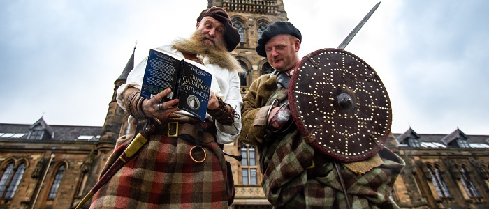 Two reenactors in period costume study a history text standing outside the University's historic Gilbert Scott Building.