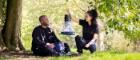 One male and one female sat on the floor outdoors surrounded by trees and grass, holding a small mosquito net. 