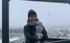 A woman stands before a snowy scene with a wind propellor in the background. She is bundled up in a hat and scarf.