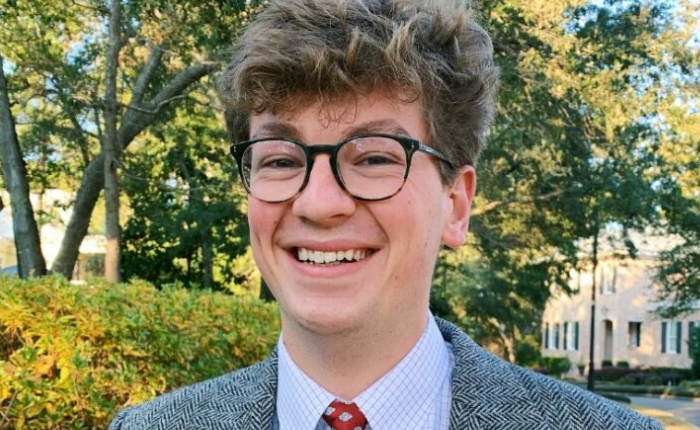 A young man in a grey suit smiles at the camera. Behind him are the green and yellow leaves of trees and a hedge