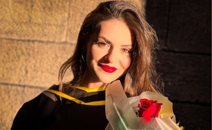 A woman in a graduation gown smiles at a camera at golden hour, holding a bouquet