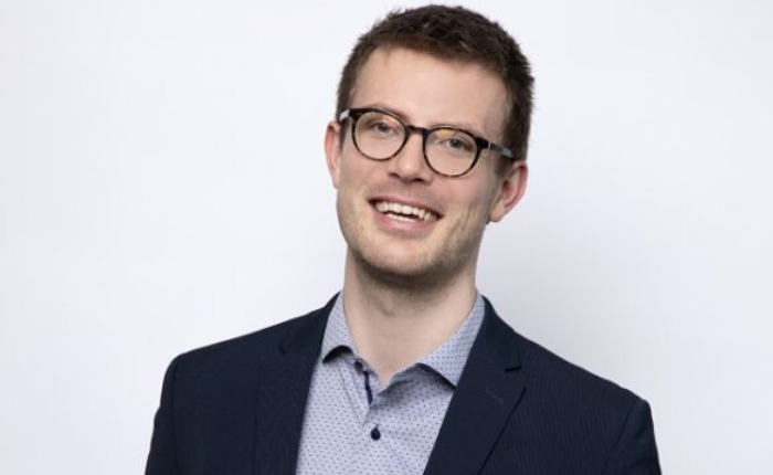 A man wearing glasses smiles before a pale grey background