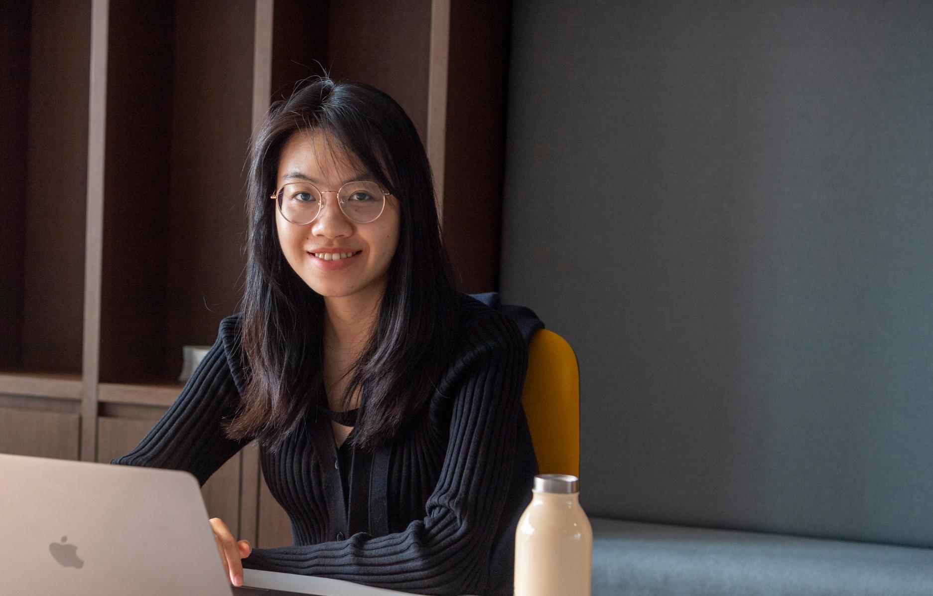 A student with long hair, wearing glasses, smiles while sitting at a desk on her laptop