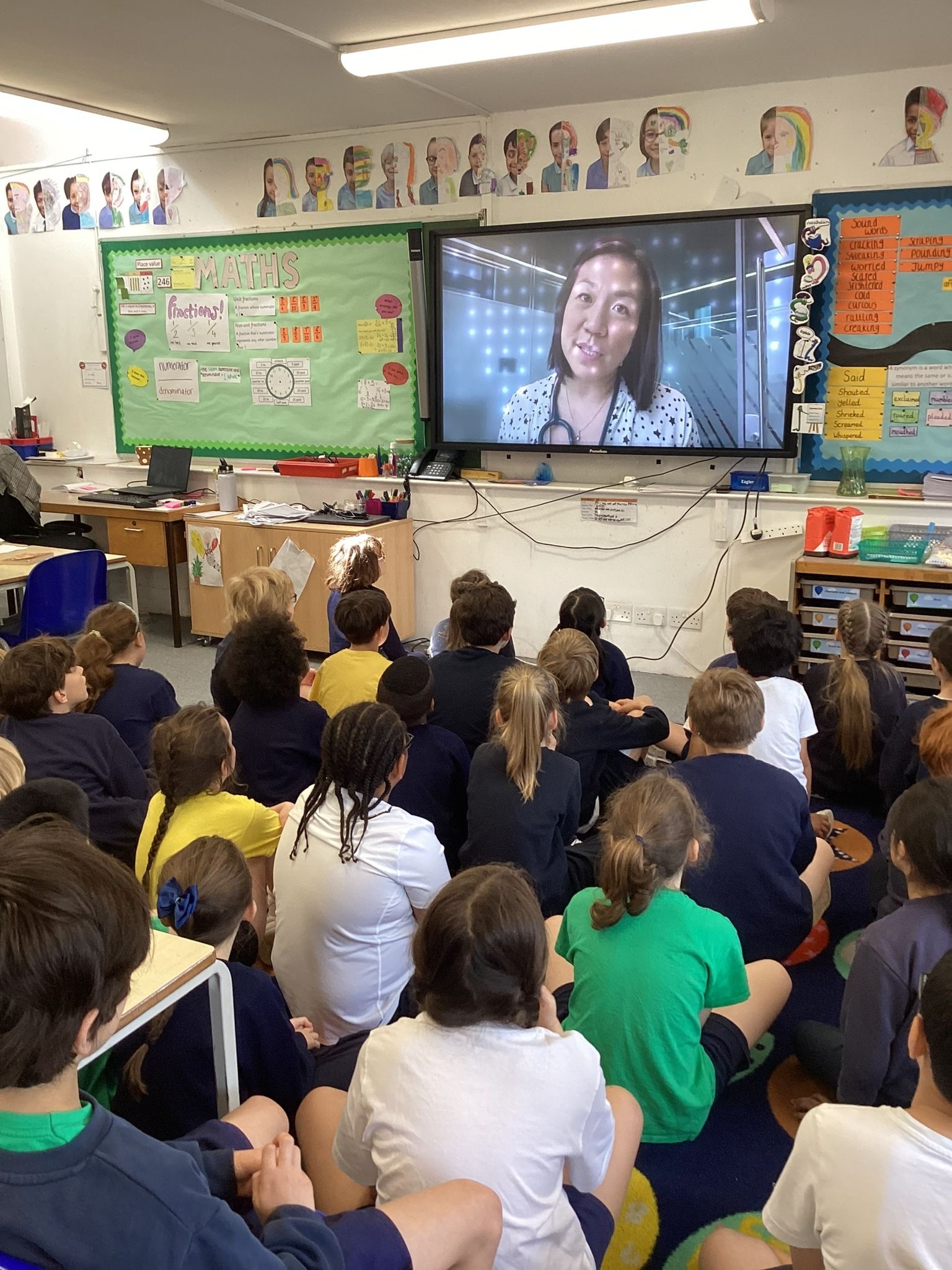 Antonia Ho on television screen in front of school pupils who are all sat on the floor watching intently