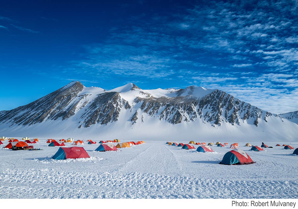 Camp in Antarctica: colourful tents on snow, with snowy cliffs in the background under a blue sky.