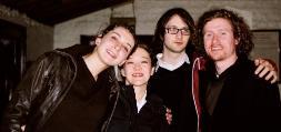 photograph of interior: a group of four young white students huddled together, smiling at the camera. background is evening outside