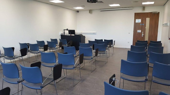 Flat floored teaching room with rows of tablet chairs, projector, screen, visualiser, and PC