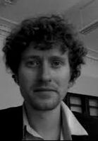 black-and-white photo of Robert Cowan: headshot of a white young man with big curly hair and short facial hair, in a white open collared shirt under a black blazer. Background seems to be that of a classroom