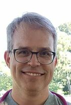 photo of Glen Pettigrove: toght headshot of older man with elegantly swept grey hair and big round glasses, with a wide smile with dimples. Backdrop is lush outdoors