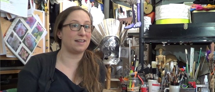 photo: Interior of an artist's studio with supplies and a wooden desk. Foreground - a woman with brown long hair, glasses and dressed in black addresses the viewer