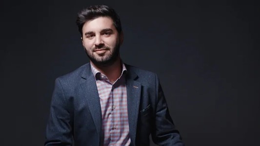 photo of a professional man in his 30s, with dark hair and short beard, in a blue suit and checkered shirt