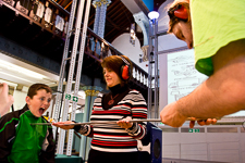 Photograph of a scientist demonstrating an experiment involving planks of wood whilst wearing sound defenders. A child is next to her yelling at the plank of wood. 