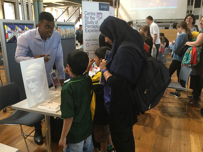 Photograph of a GSF volunteer explaining an Albert Einstein illusion image to a woman and 2 children. 