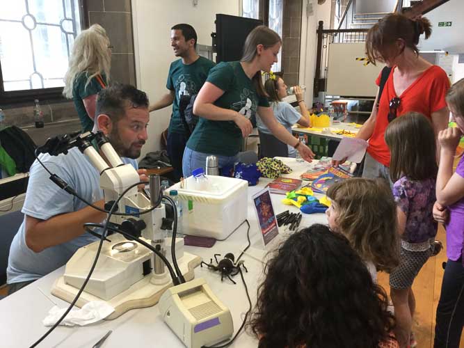 Photograph showing a GSF volunteer in front of a microscope with several children listening to the explanation. In the background another vounteer is passing out magazines. 