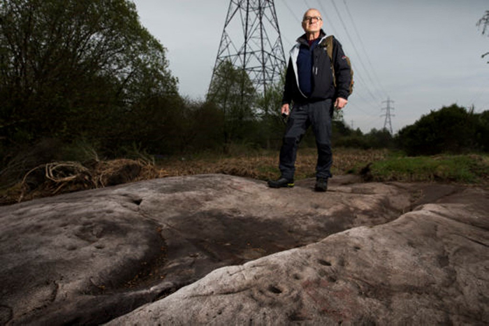 Gordon Morrison, an older gentleman, stands on one of the Auchnacraig rock-art panels at Faifley.