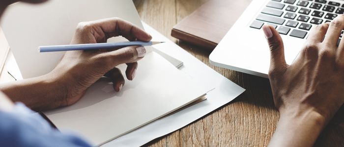 Photo of person using a laptop and writing in a notepad