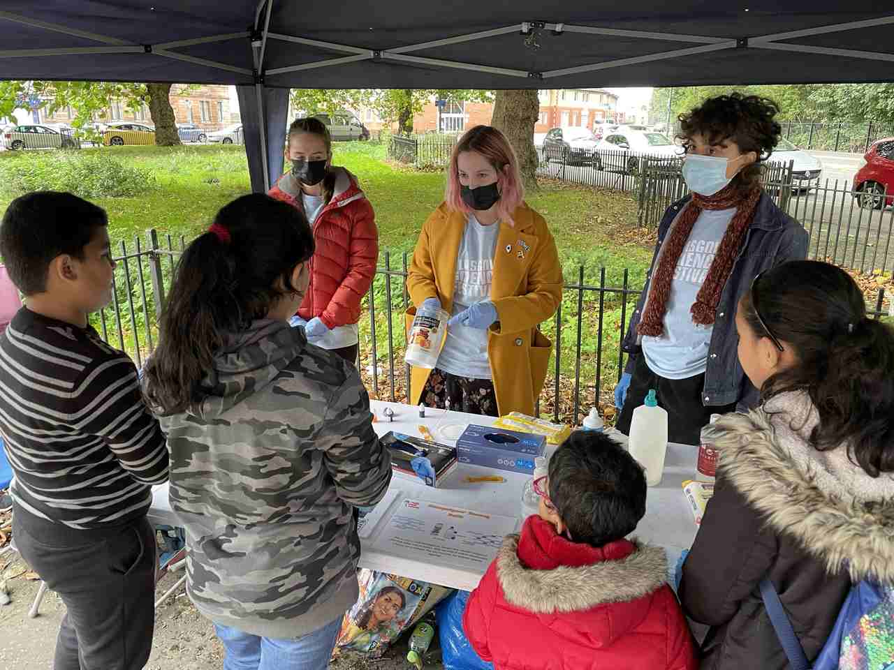 The picture was taken outdoors and inside a gazebo. Here three GSF staff members are doing a demonstration of an activity to a group of four kids. Inbetween GSF staff members and children, there is a table where they can engage in the activity being showed to them.