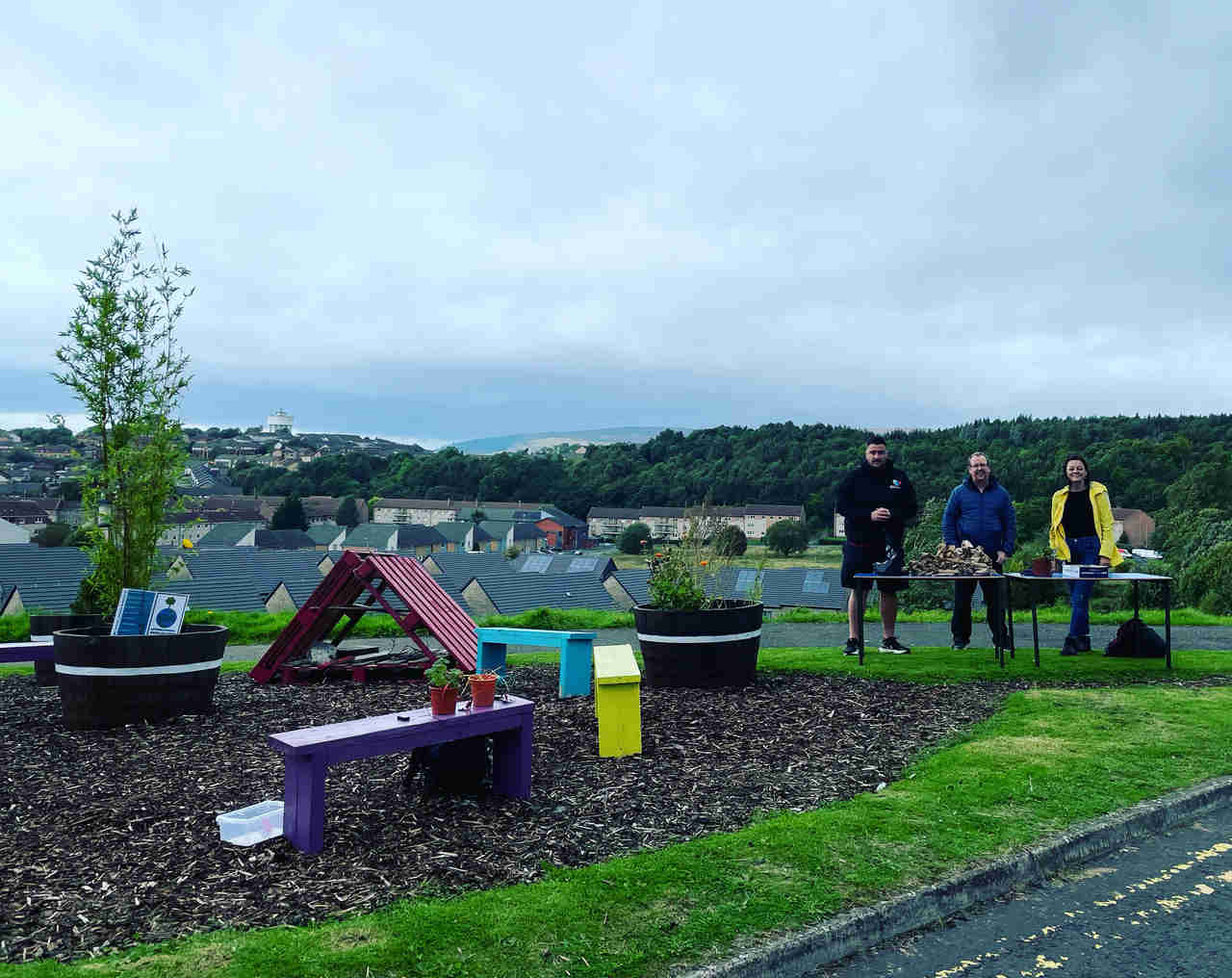 A flowerbed is in the picture. There is some grass with small yellow flowers one the left-hand side and soil on the right-hand side. The flowerbed has a wooden fence. Two watering cans (a red and a green one) are outside the flowerbed. 