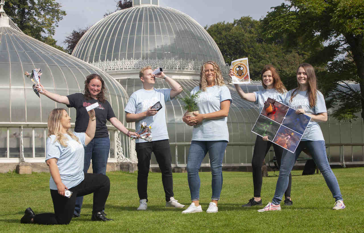 A group of adults stood in front of a large glass greenhouse are holding various science related items. From left to right these are a spectroscope, recycled paper flowers, a plant in pot, a bee poster and a tic poster. 