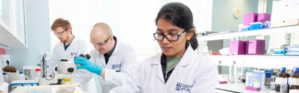 An image showing three scientists in UofG lab coats stood at lab bench in a row conducting different research