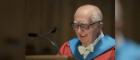 Older man wearing glasses and robes standing at lectern 